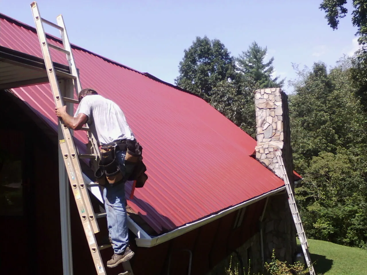 Roofer on a ladder beside a red metal roof and stone chimney — Soffit & Fascia Repair by Roofing Brothers in Lynnfield